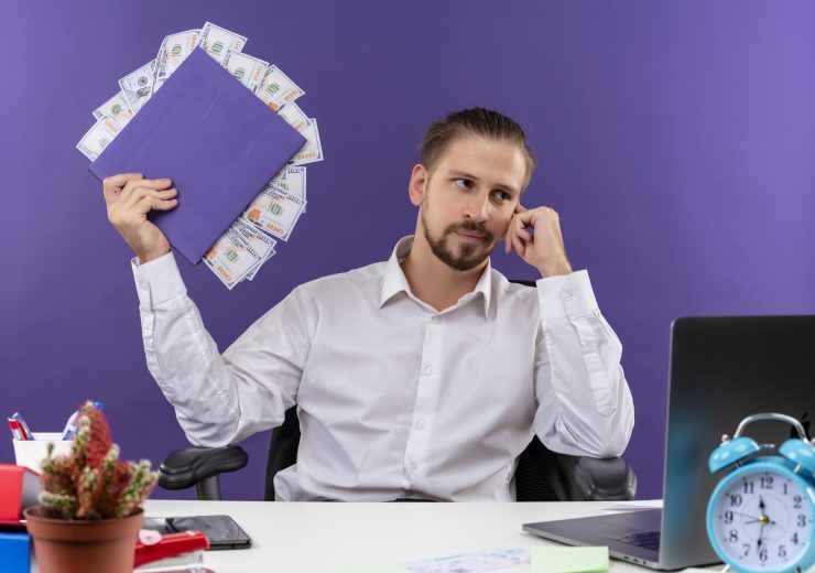 handsome businessman in white shirt holding folder with cash looking aside with pensive expression sitting at the table in offise over purple background